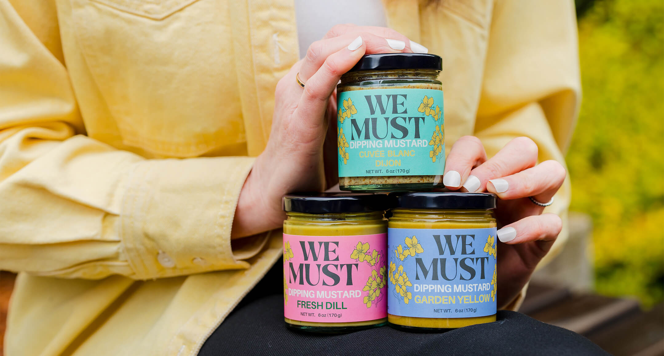 Three jars of We Must mustard including dijon, garden yellow, and fresh dill flavors.