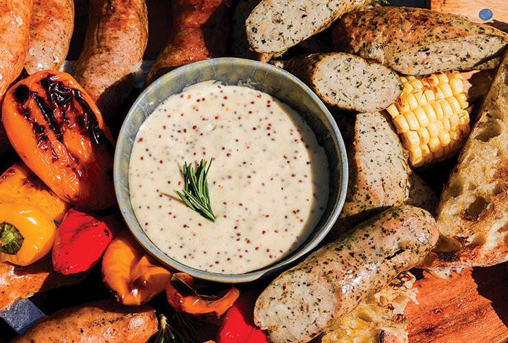 A bowl of dijonnaise plated with grilled sausages, corn and mini peppers